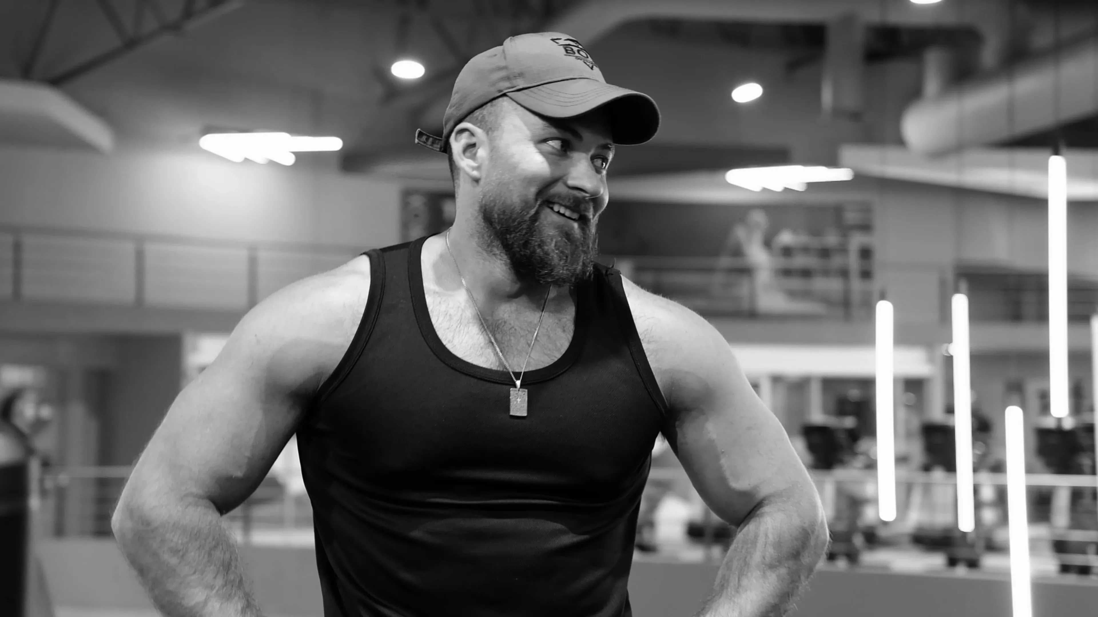 Coach Ronnie smiling in the gym, hands on hips, wearing a Bodies by Ronnie cap and vest.